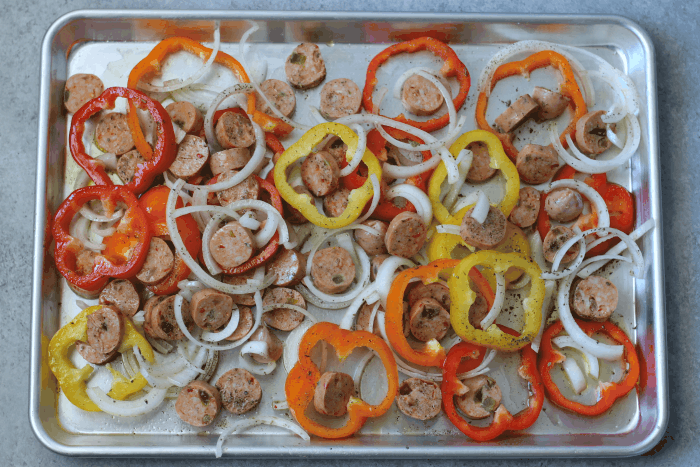 Sheet pan filled with slices of chicken sausage, slices of onion, and thin slices of orange, red, and yellow bell peppers.