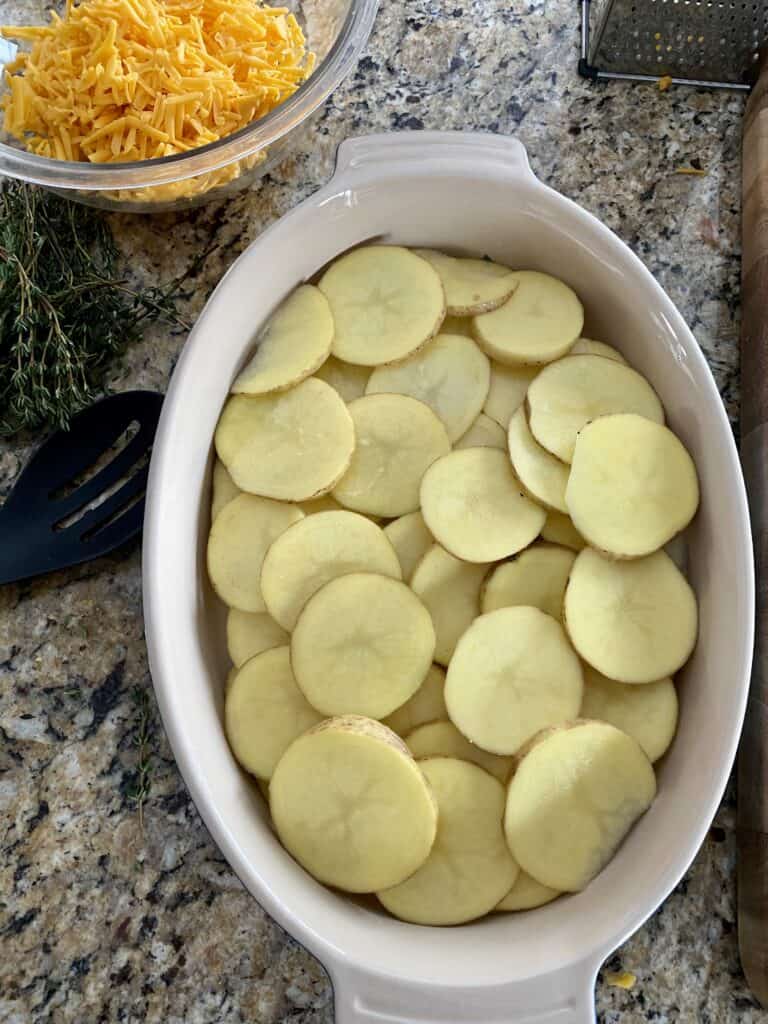 Thinly sliced round potatoes in an oval baking dish with a bowl of shredded cheese in the background.