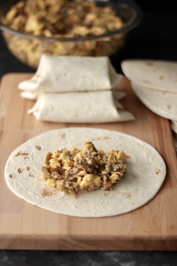 Tortilla with a scoop of breakfast burrito filling in the center and rolled breakfast tacos in the background