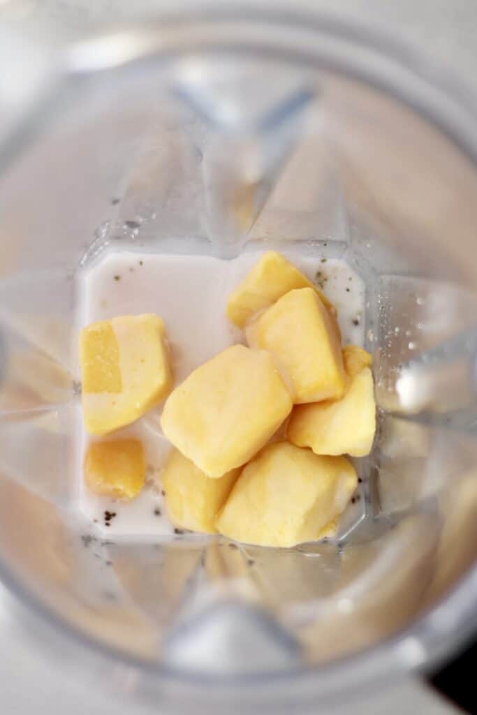 Chunks of frozen mango at the bottom of a blender container with almond milk