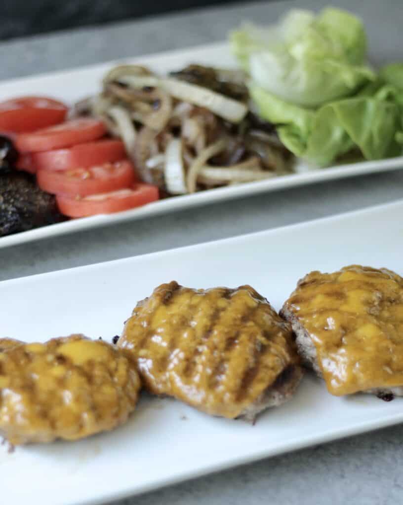 Burger patties covered in cheese next to a plate full of vegetable burger toppings