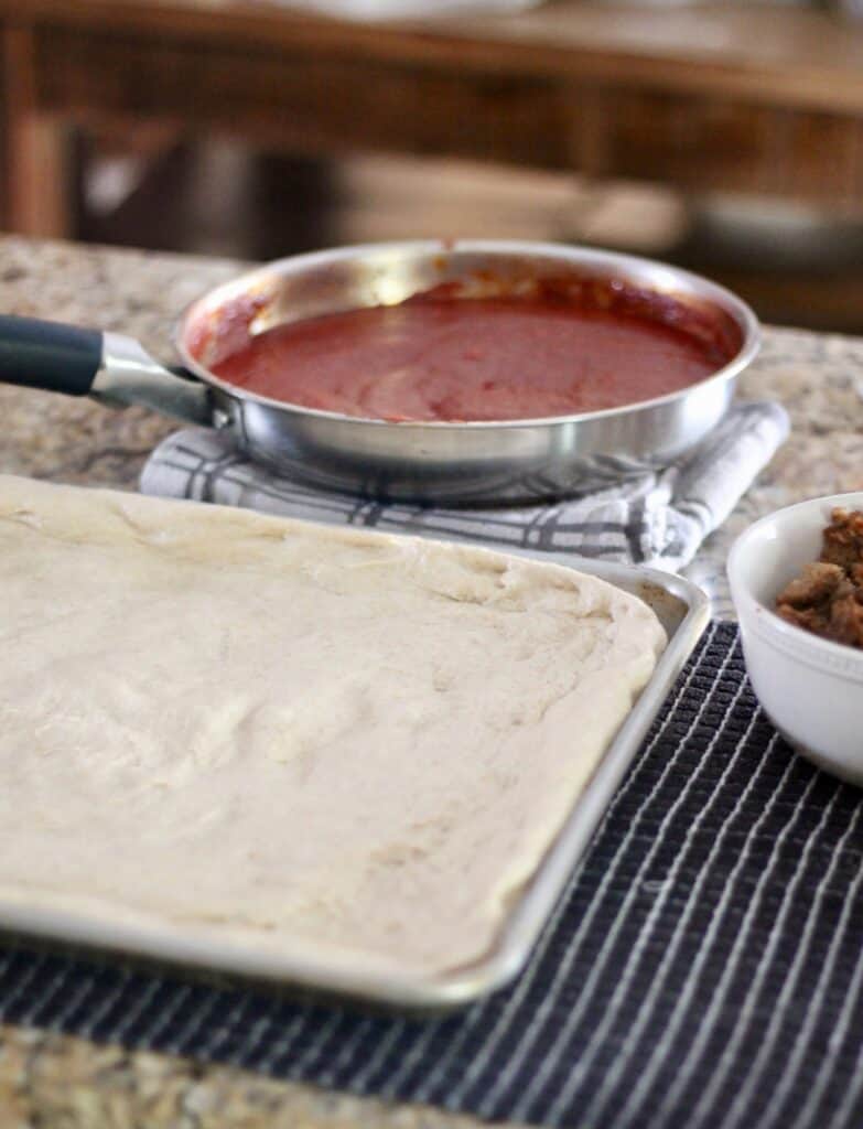 Pizza dough on a sheet pan with homemade pizza sauce in the background