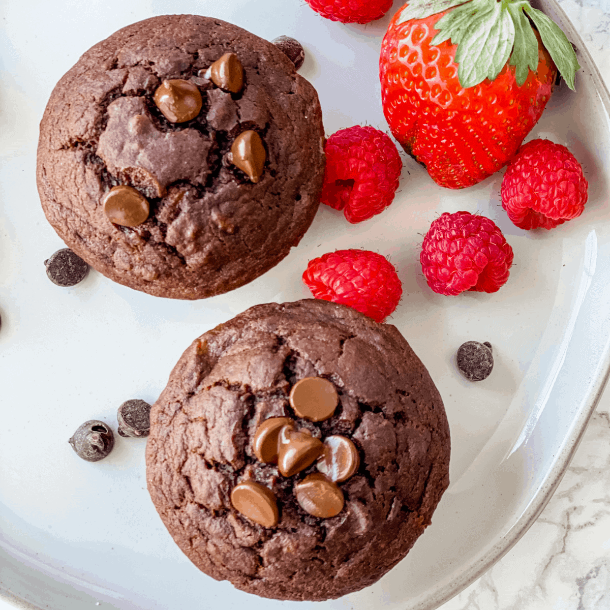 two chocolate banana muffins on plate with fresh raspberries and strawberries.