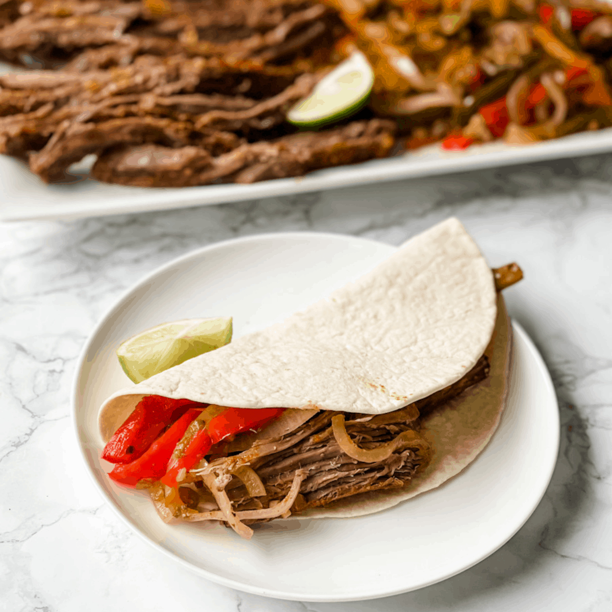 Steak fajita in a flour tortilla with a platter of sliced steak, onions, and peppers in the background.