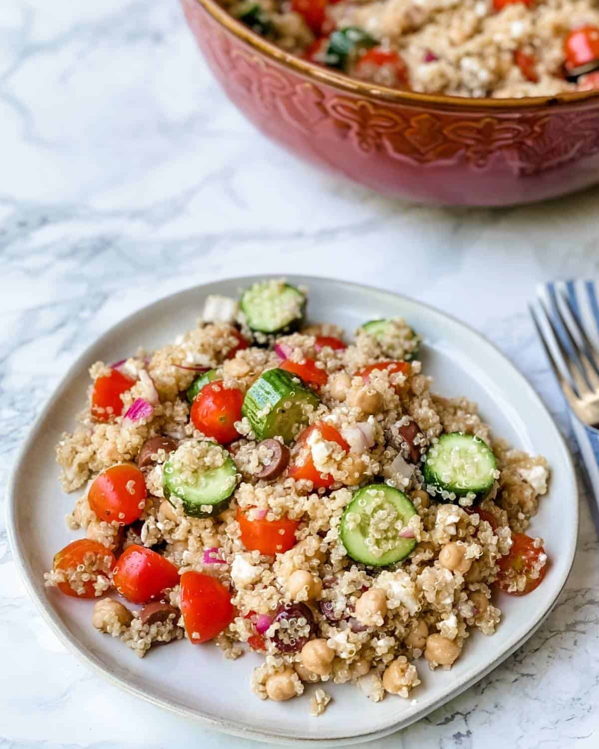 a plate of quinoa salad with feta and olives and vegetables next to a large salad bowl