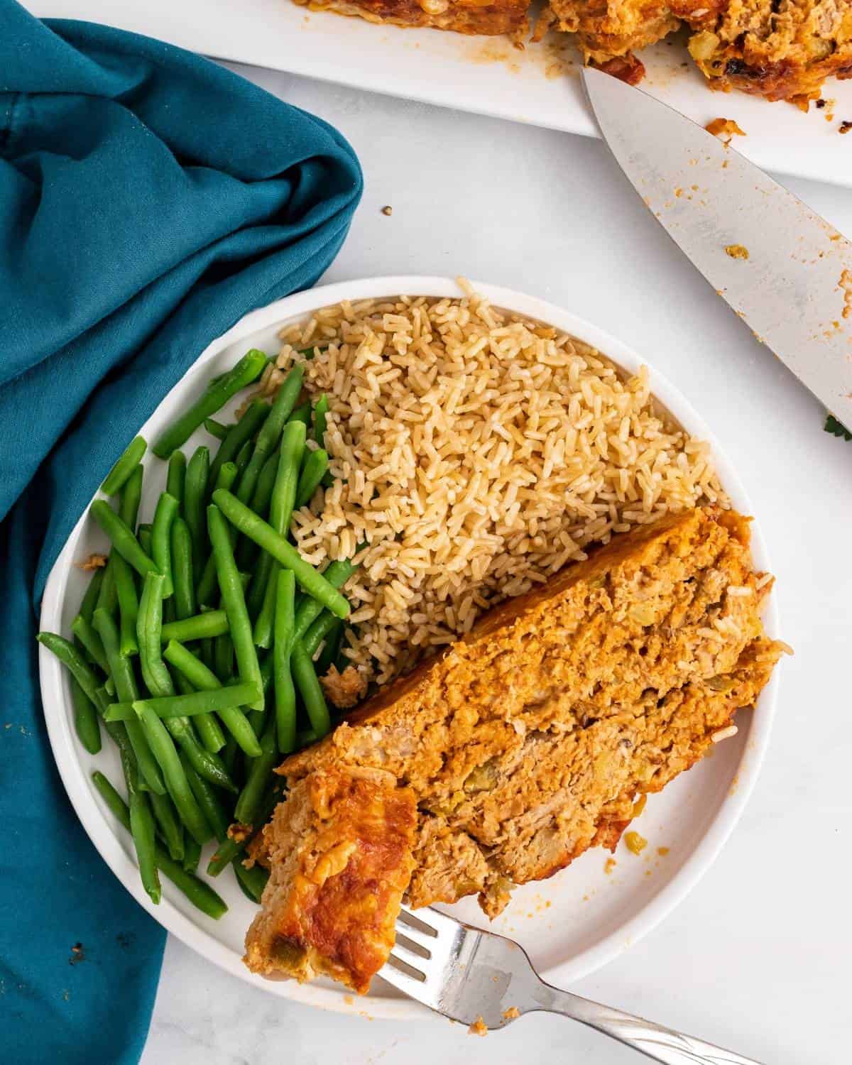 One slice of meatloaf on a plate with green beans and brown rice.