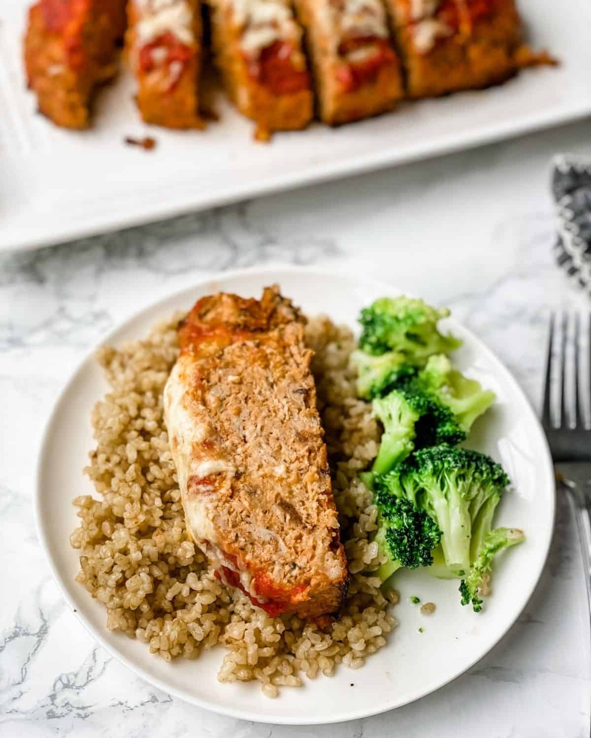 Italian meatloaf on a plate with rice and broccoli with a platter of sliced meatloaf in the background.