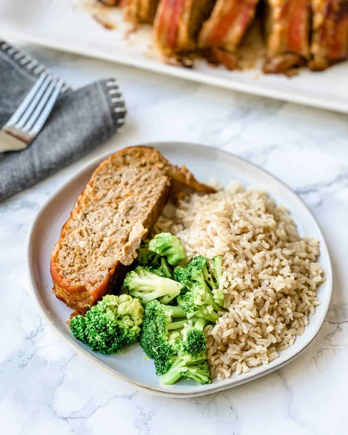 a piece of bacon cheeseburger meatloaf on a plate with broccoli and rice.