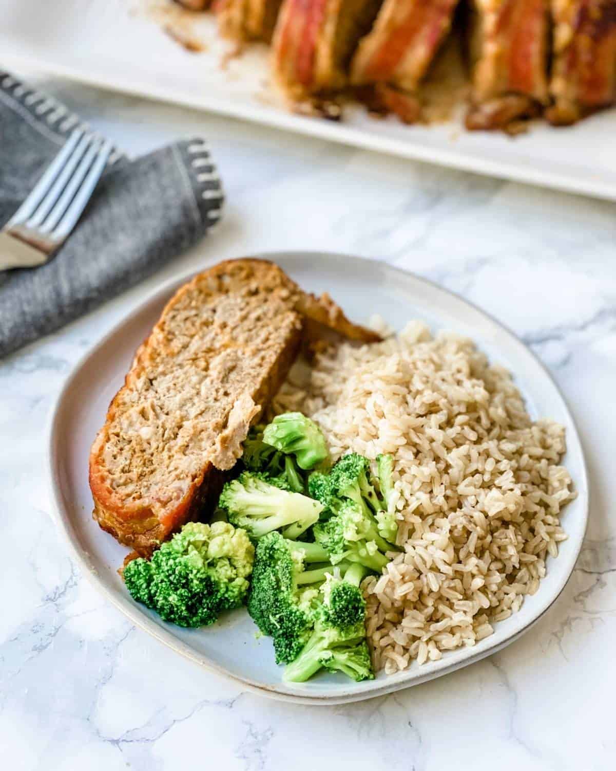 A piece of bacon cheeseburger patty is placed on a plate with broccoli and rice on it.