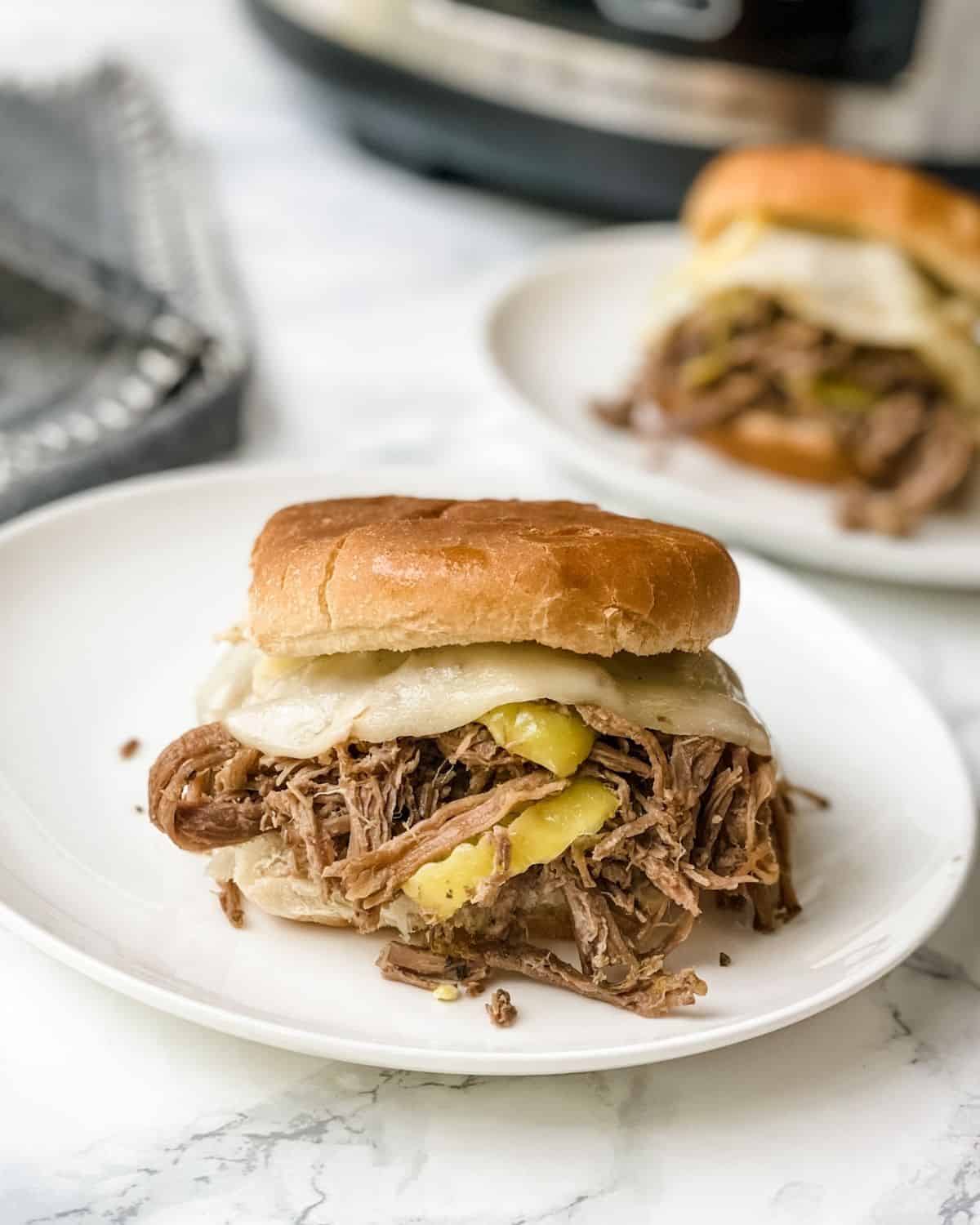 A closeup picture of the italian shredded beef on a roll with cheese. There's another sandwich behind it with the slow cooker next to it.