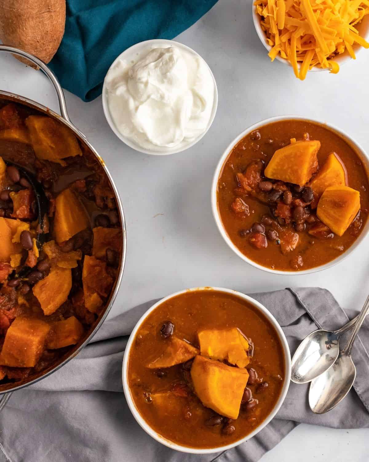 The sweet potato and black bean chili are placed in two bowls with two spoons next to them. There is a pot of chili soup next to the bowl.