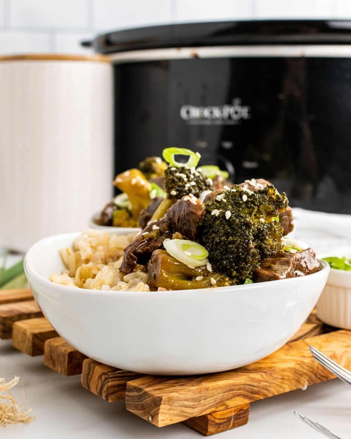 A closeup picture of beef and broccoli in a white bowl with a crockpot in the background.