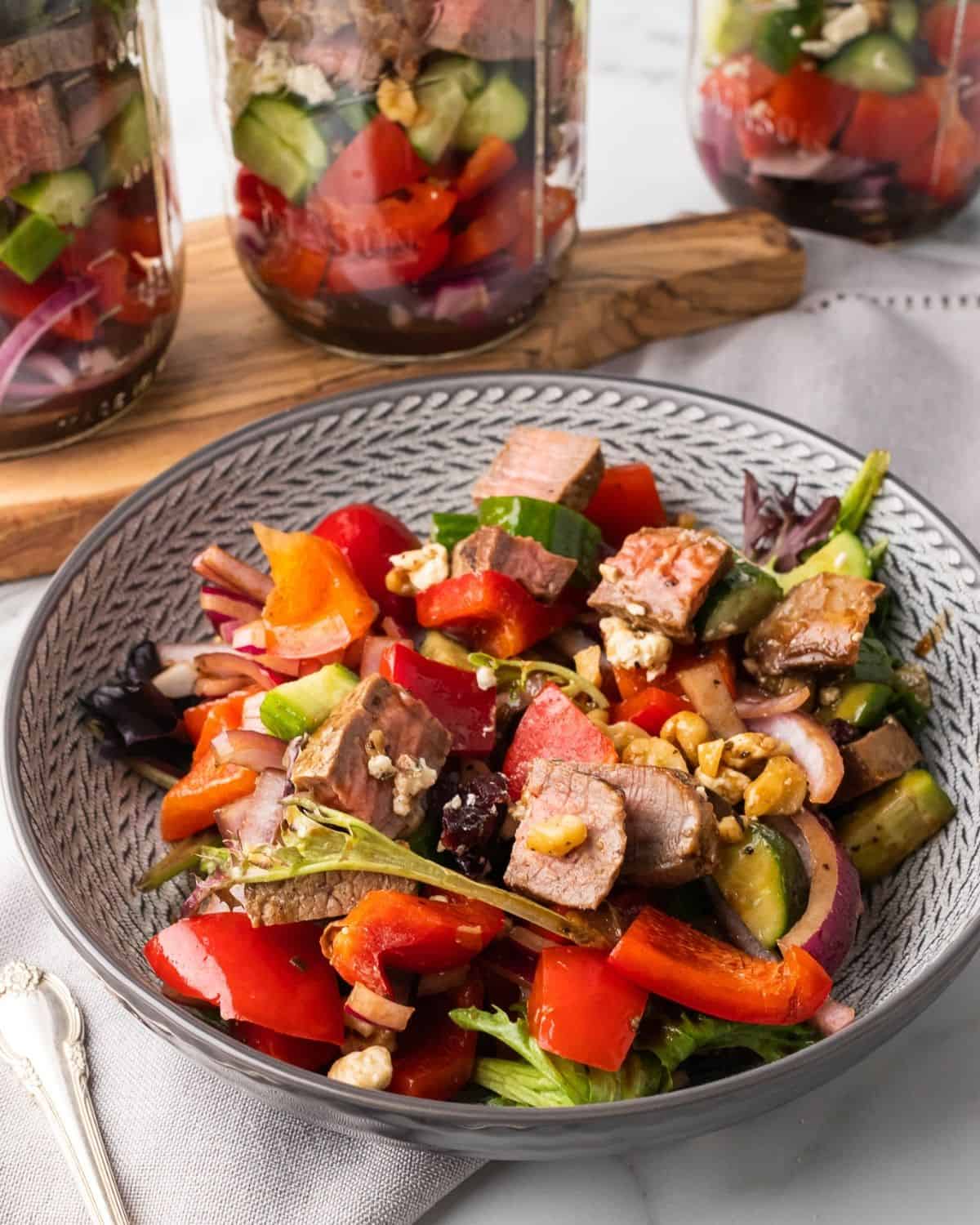 steak salad in a large grey bowl with mason jar salads in the background.