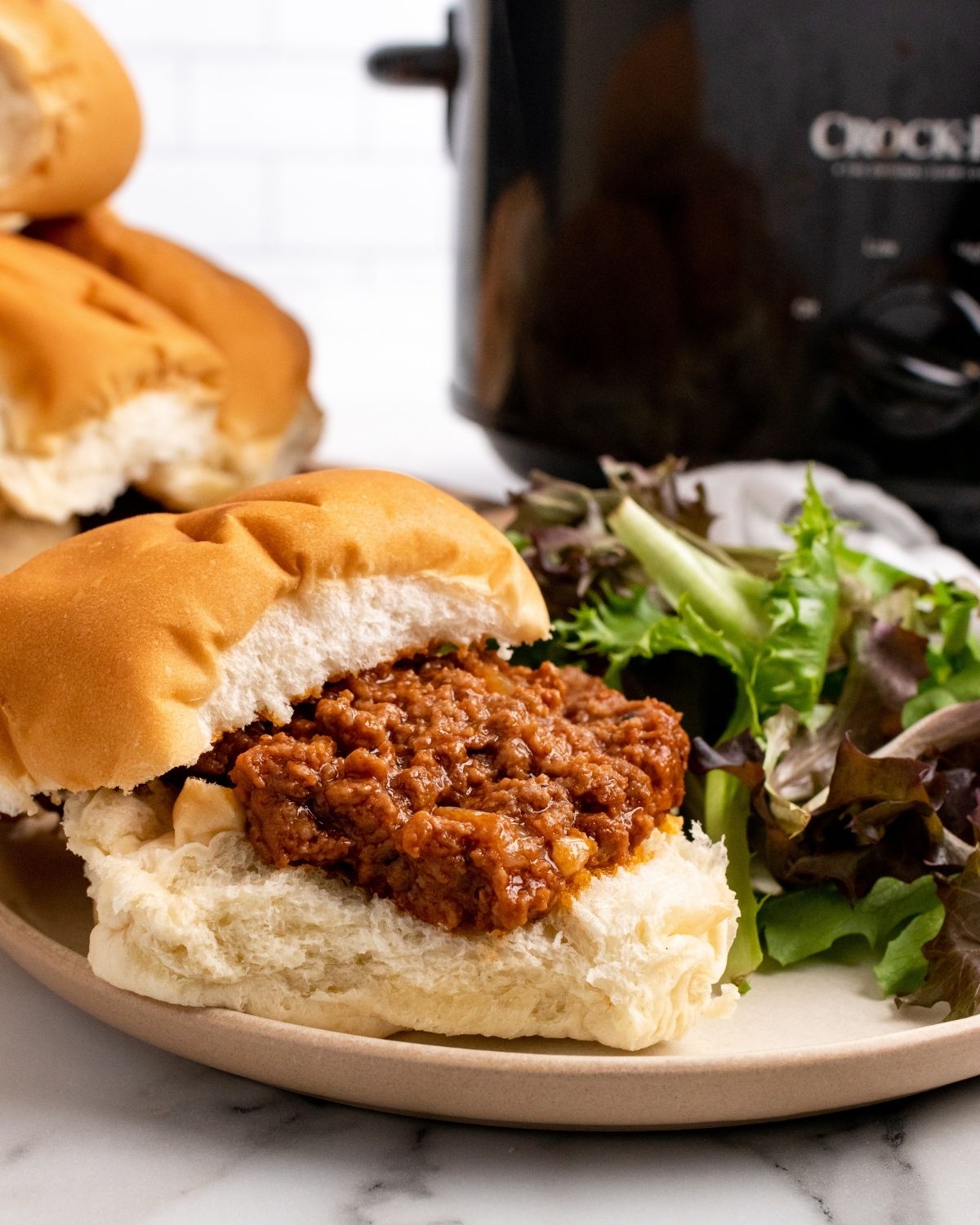 sloppy Joe on a bun on plate with salad and the Crockpot in the background.