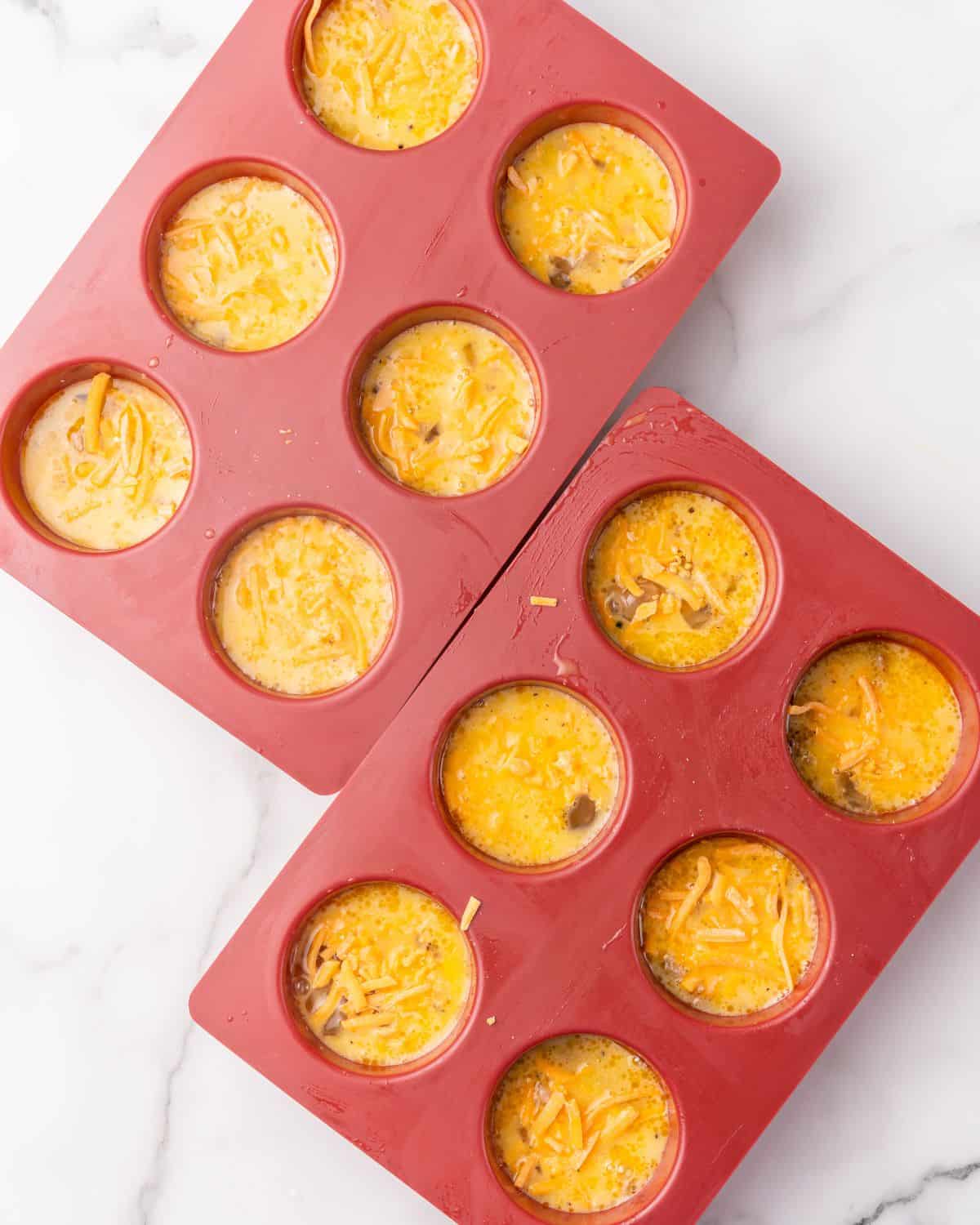 egg mixture in the muffin pans. Cheddar cheese is sprinkled on top of the egg cups.